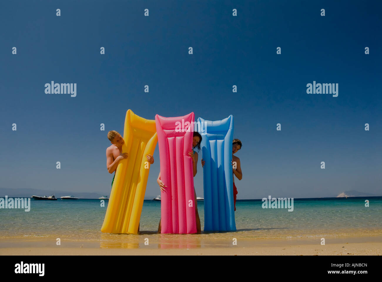 Three people looking out from behind colourful lilos inflatable airbeds sandy beach blue sea and sky Mediterranean Greece Stock Photo