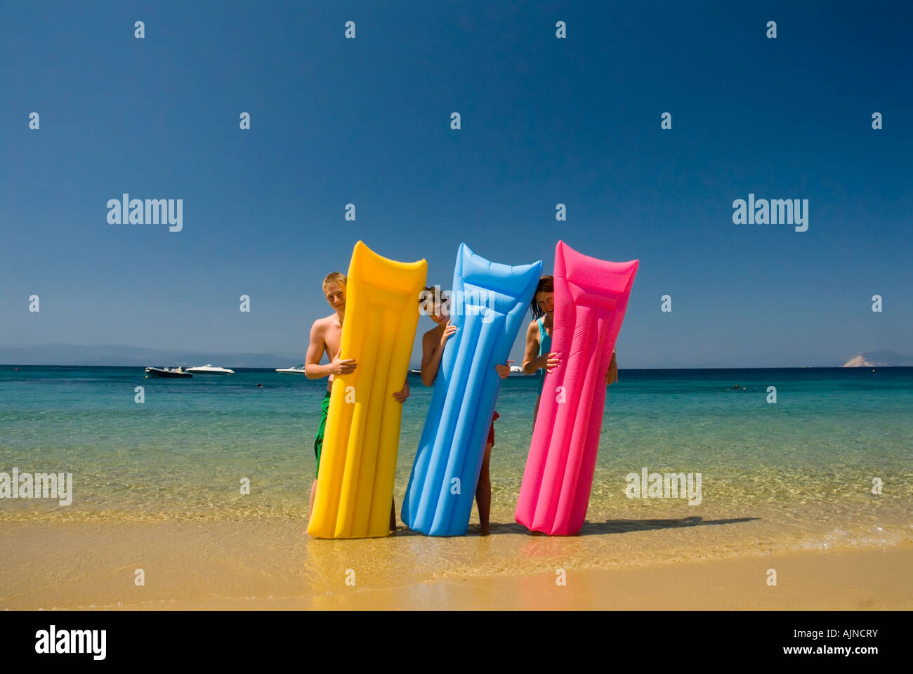 Three people looking out from behind colourful li los inflatable airbeds sandy beach blue sea and sky Mediterranean Greece Stock Photo