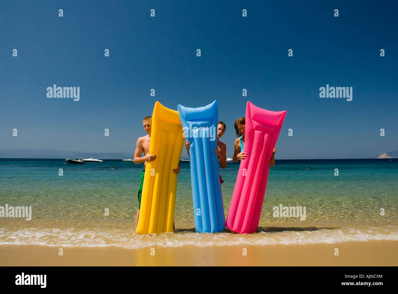 Three people looking out from behind colourful li-los inflatable airbeds sandy beach blue sea and sky Mediterranean Greece Stock Photo