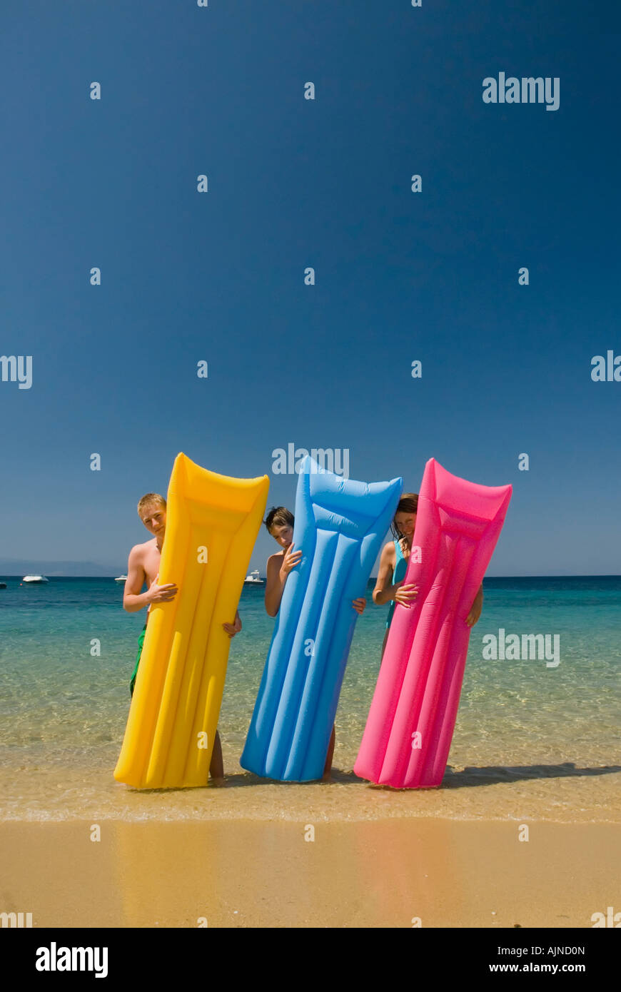 Three people looking out from behind colourful li-los inflatable airbeds sandy beach blue sea and sky Mediterranean Greece Stock Photo