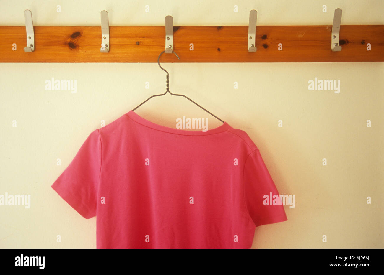 Pink T-shirt on wire hanger hanging from metal hook in row of clothes hooks mounted on wood on cream wall Stock Photo