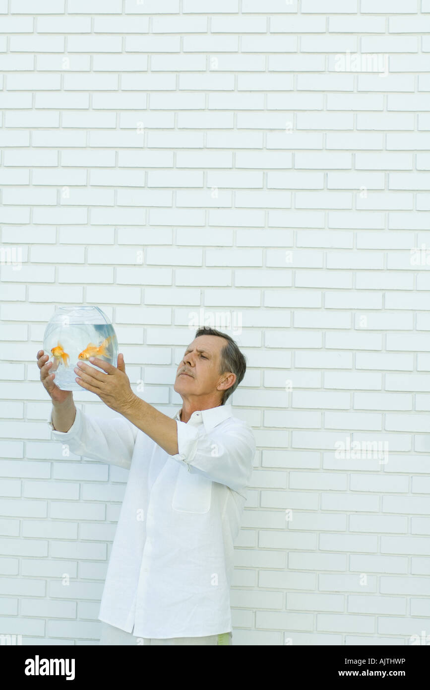 Man holding up goldfish bowl, standing in front of white brick wall, waist up Stock Photo