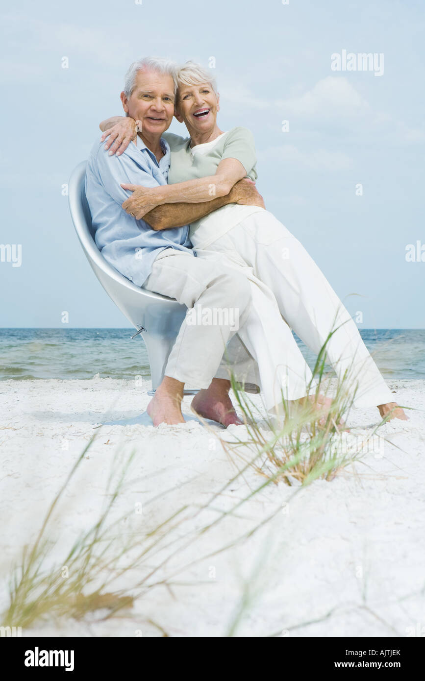 Senior couple sitting in chair together on beach, embracing, woman sitting on man's lap, both smiling at camera Stock Photo
