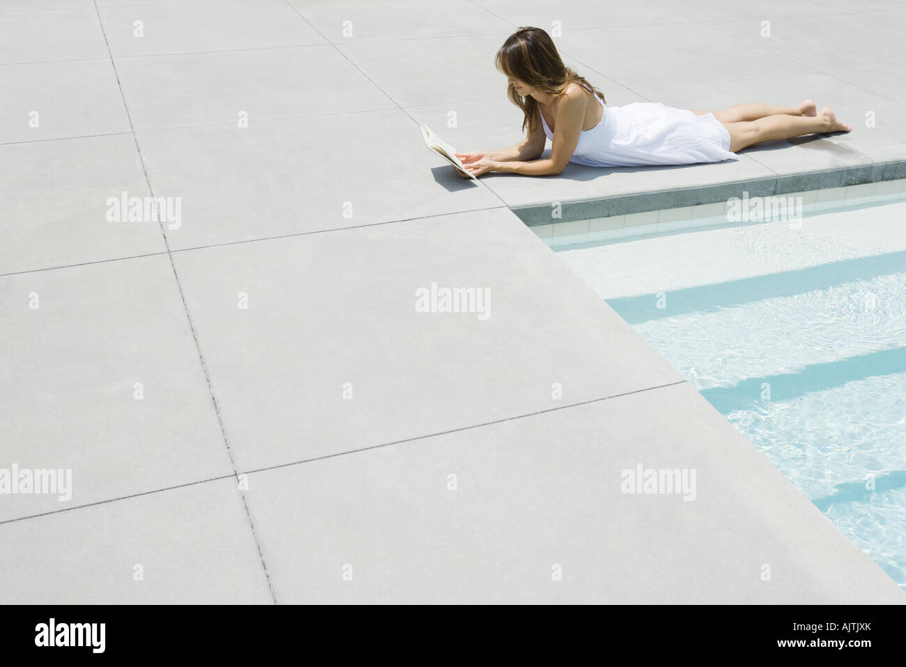 Woman in sundress lying by edge of pool, reading, full length Stock Photo