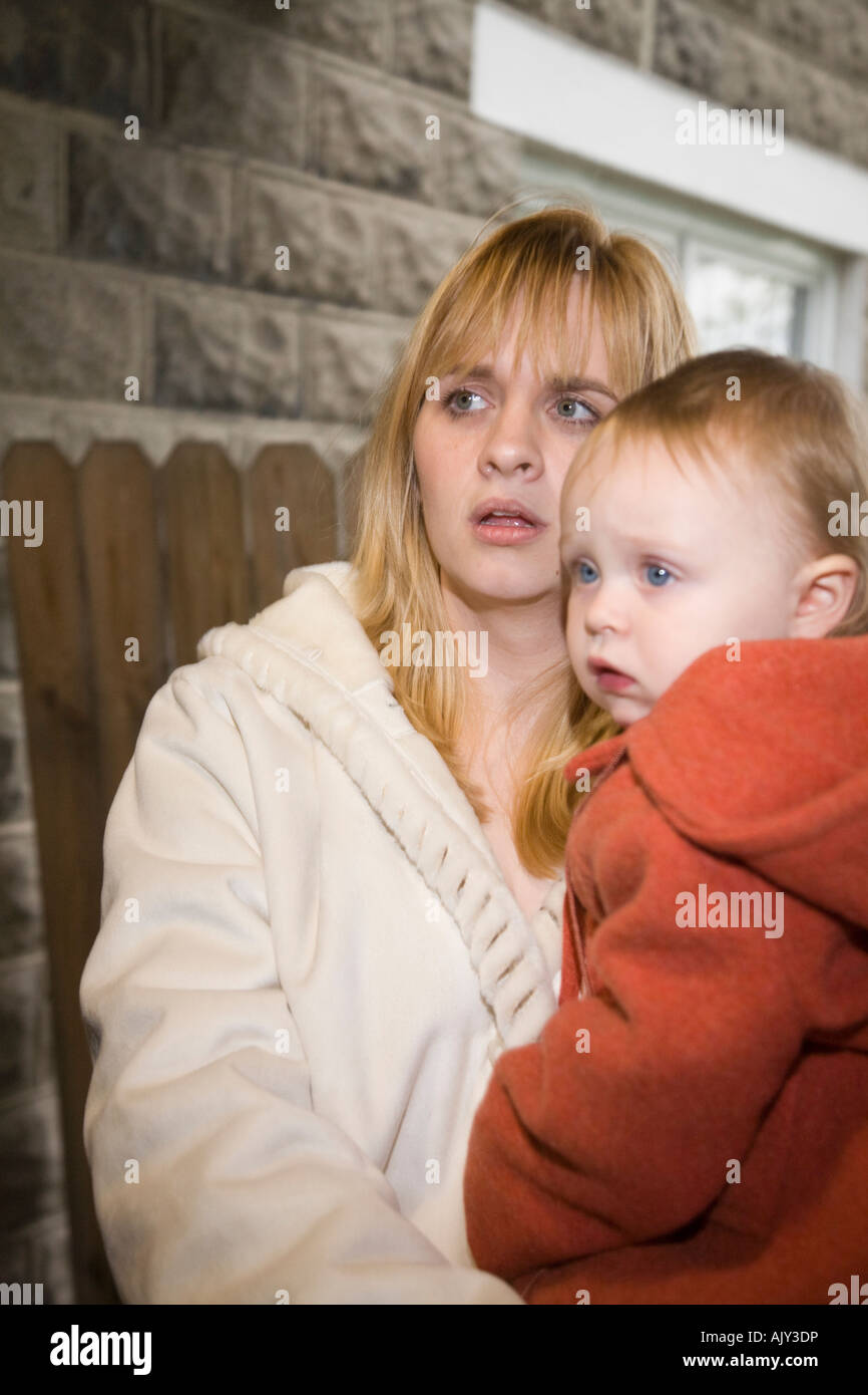Wary young mother holding her young toddler outside a dimly-lit building. Stock Photo