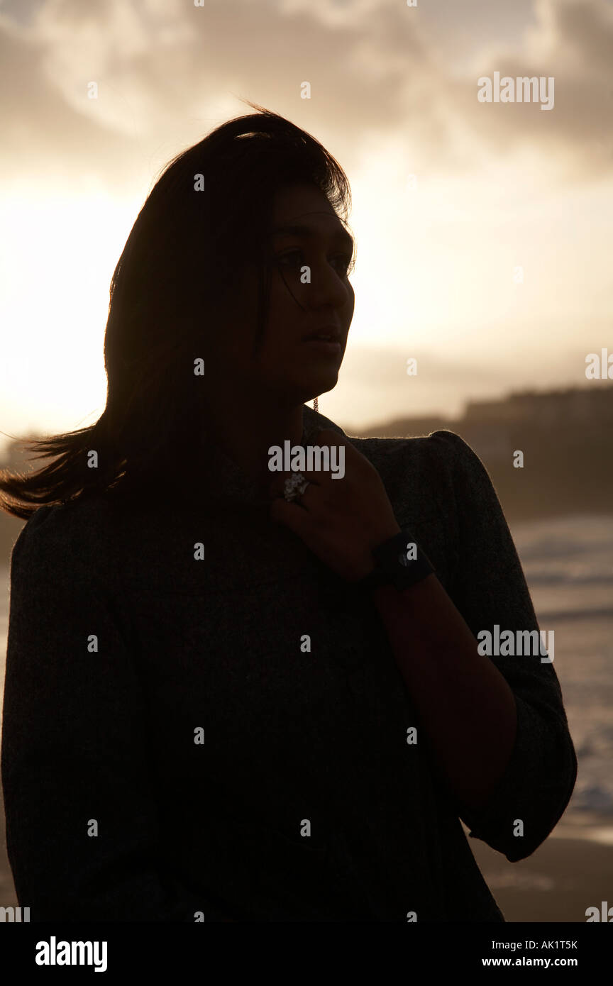 silhouette of dark haired asian female student standing on a beach at sunset looking out to sea with hand held up to her neck Stock Photo