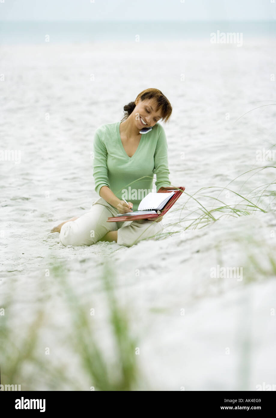 Woman sitting on sand, using cell phone and writing in agenda Stock Photo