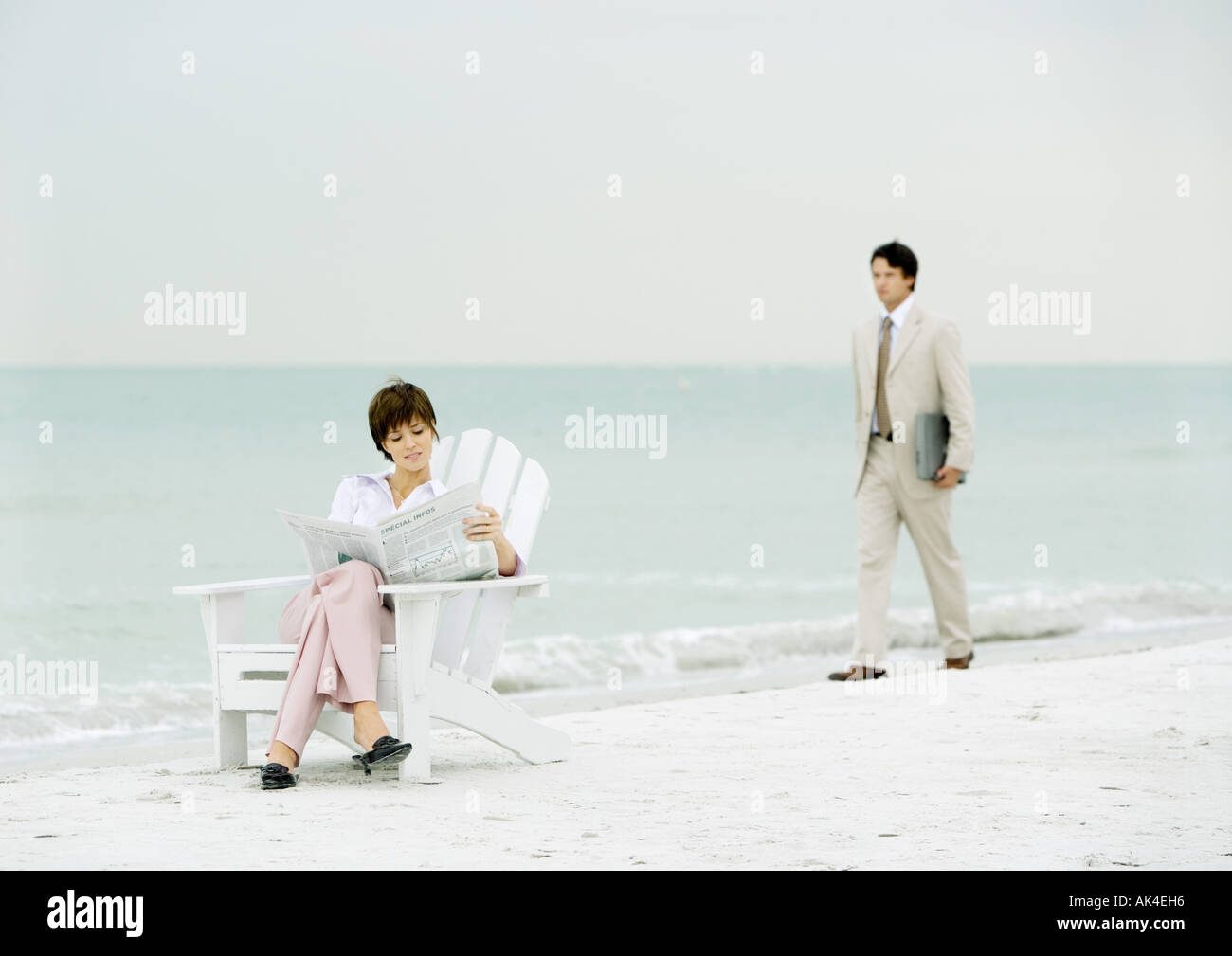 Woman sitting reading newspaper on beach as businessman carrying laptop approaches Stock Photo