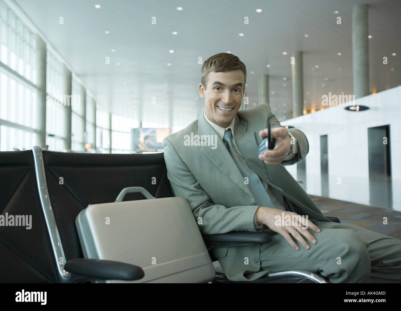 Businessman sitting in airport lounge, holding out cell phone toward camera Stock Photo