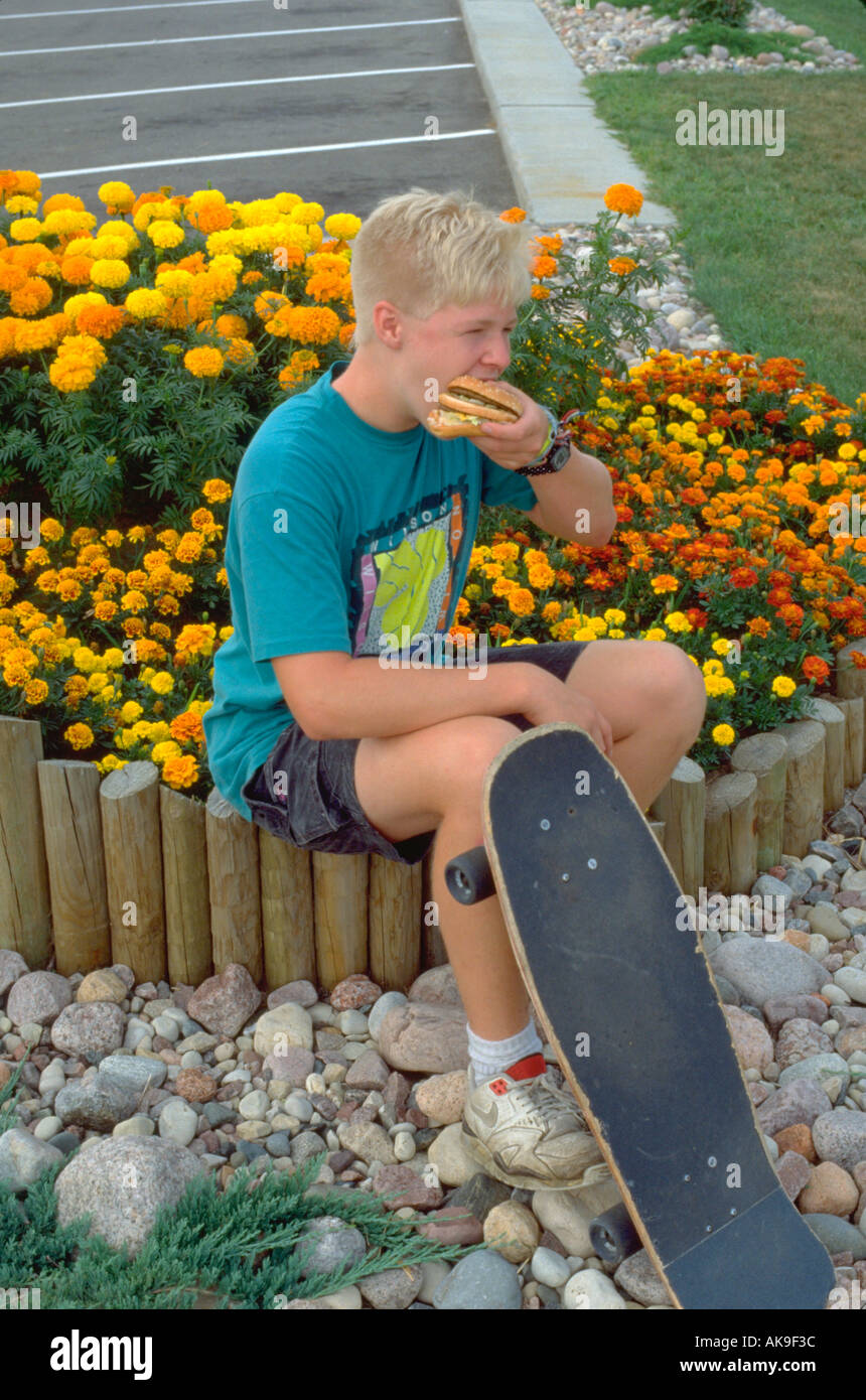 Skateboarder age 15 eating fast food hamburger by flowers. South Dakota USA Stock Photo