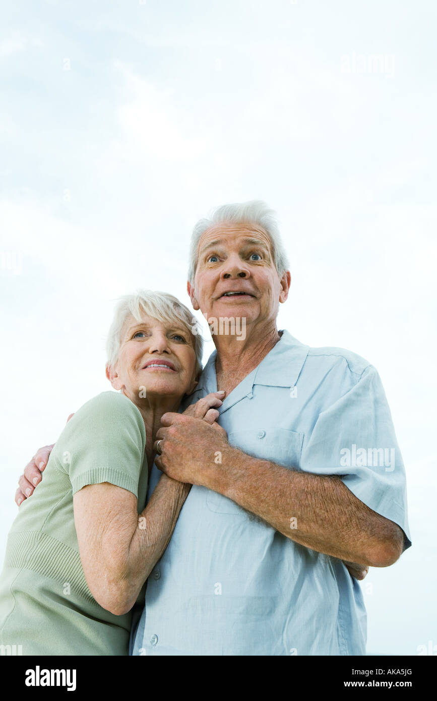 Senior couple embracing and looking away, man raising eyebrows, low angle view, waist up Stock Photo