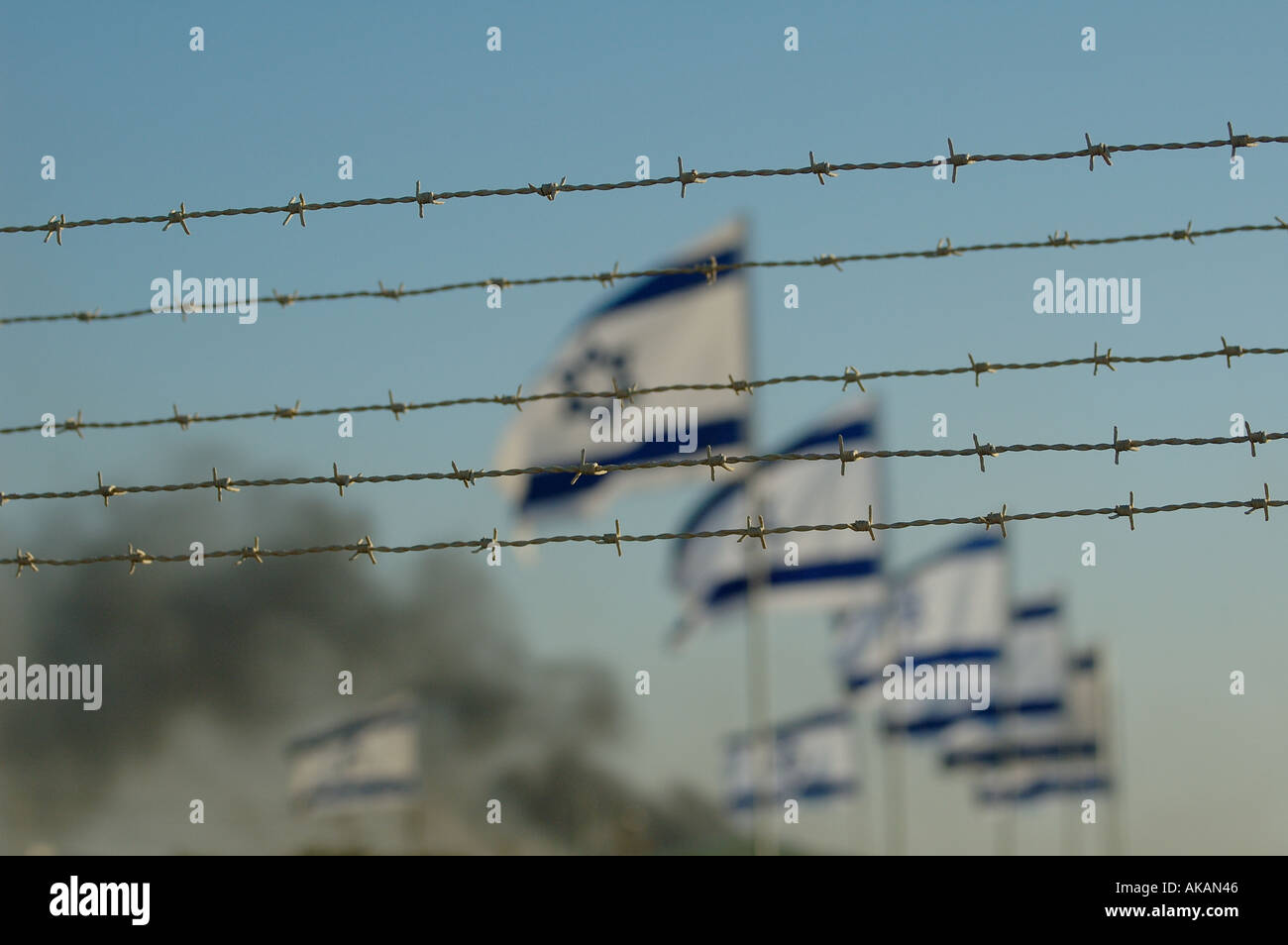 Row of Israeli flags flutter in the wind behind barbed wire fence with heavy smoke in background during Jewish settlement evacuation from Gaza strip Stock Photo