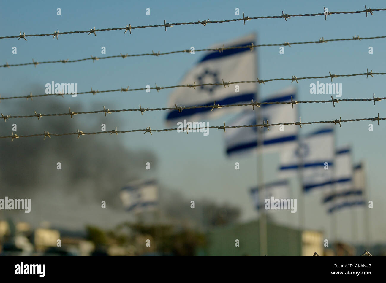 Row of Israeli flags flutter in the wind behind barbed wire fence with heavy smoke in background during Jewish settlement evacuation from Gaza strip Stock Photo