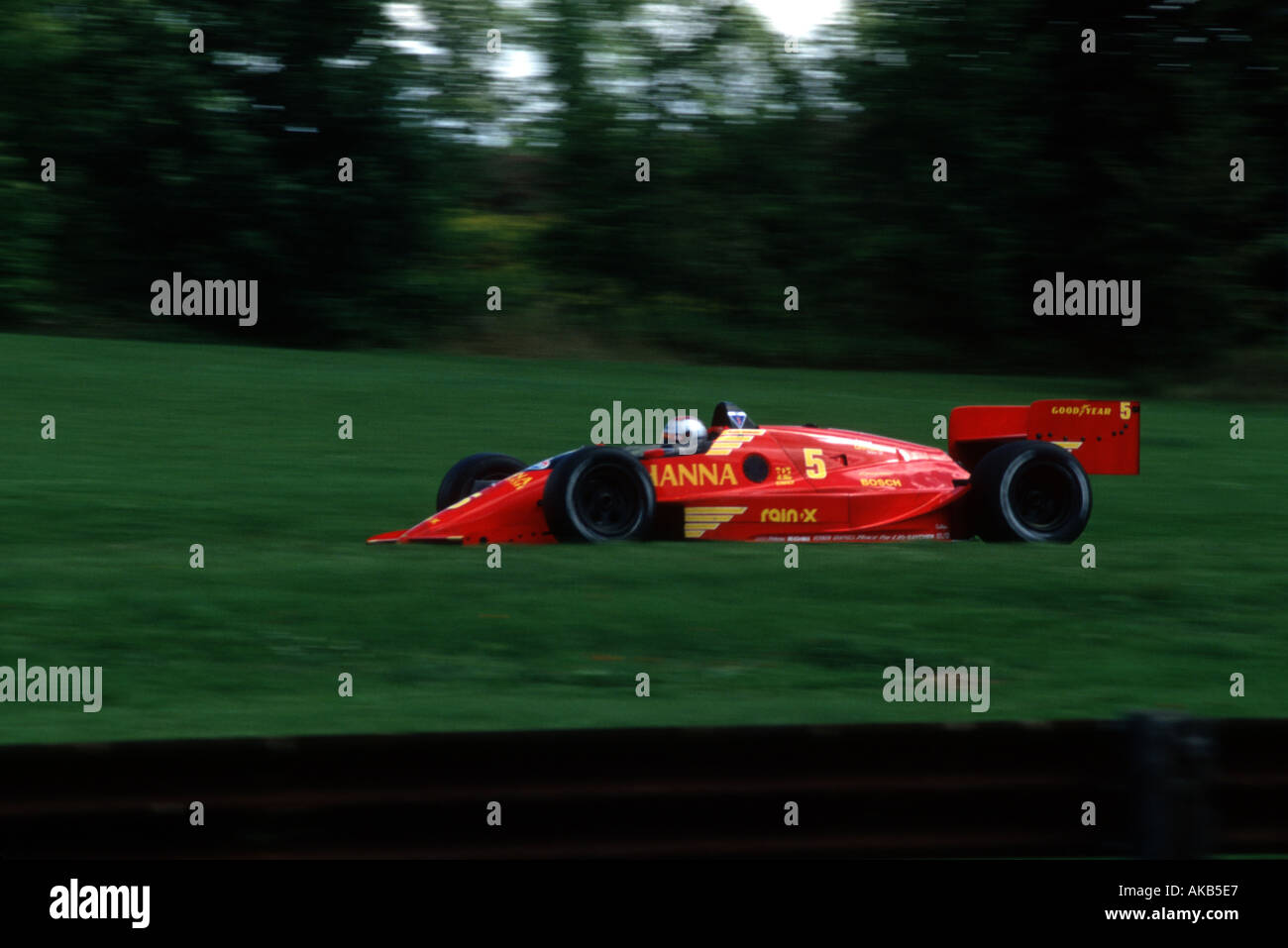 A sleek bright red race car with driver wearing a helmet speeds across a blurred green forest background Stock Photo