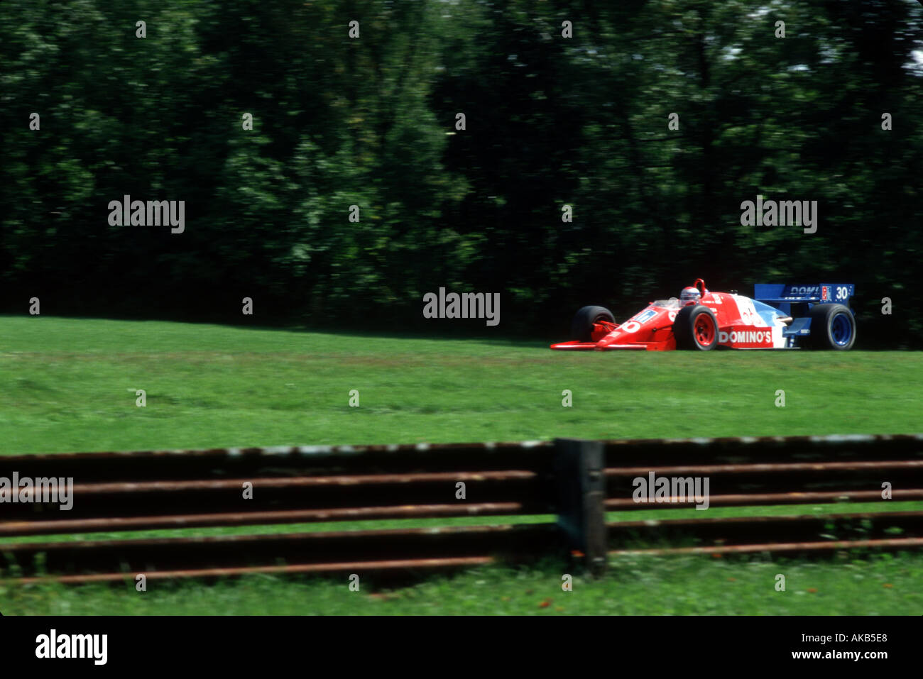 Riding low against a brilliant green grass landscape a bright red race car and driver speeds across a hill top lawn Stock Photo