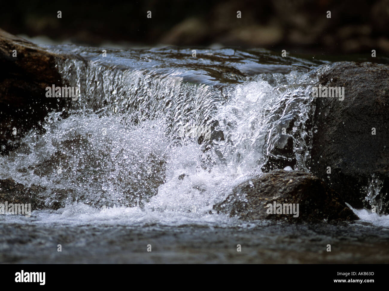 Clear cool refreshing water slips over the edge of dark granite rock and splashes forward into a close up river stream Stock Photo