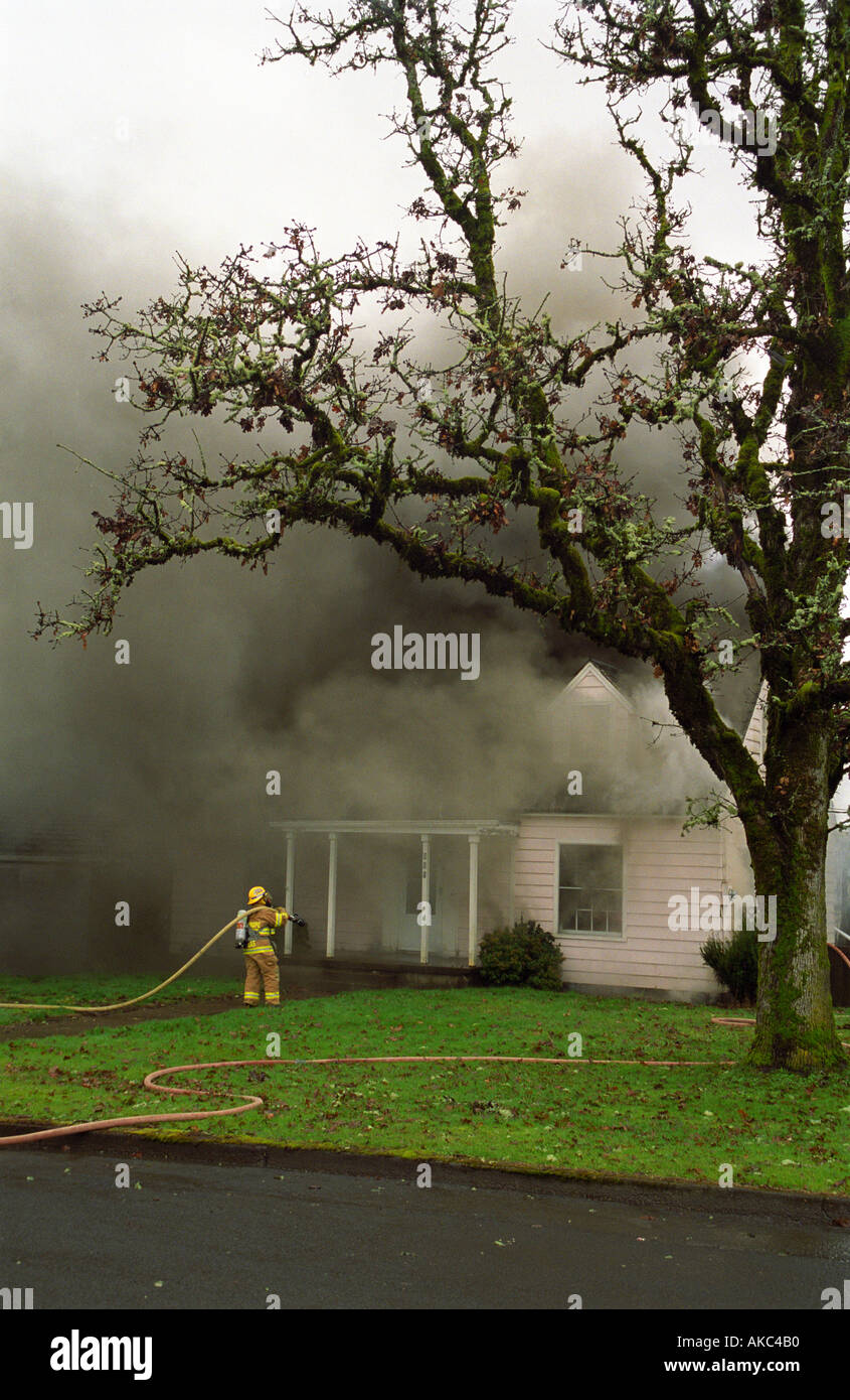 Firefighter with hose stands in front of burning smoking home Stock Photo