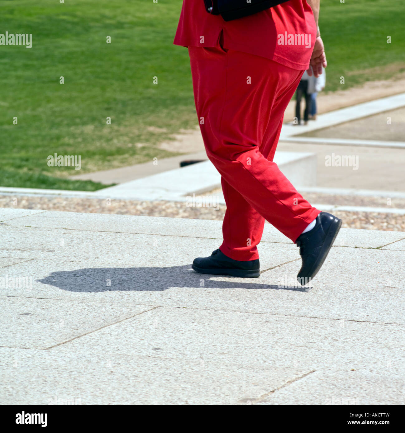 Overweight woman wearing a red pantsuit and black shoes casts a small shadow as she walks on sidewalk Stock Photo
