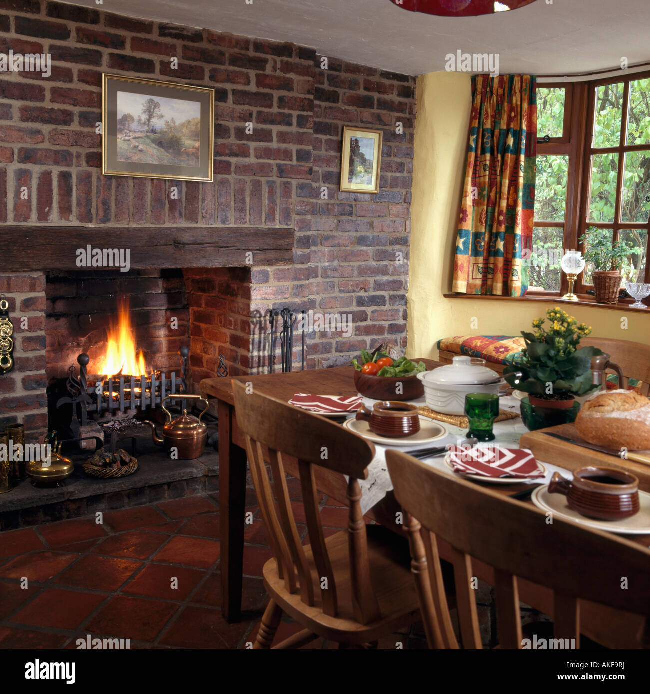 Lighted fire in fireplace in exposed brick wall in cottage dining room with table set for lunch Stock Photo