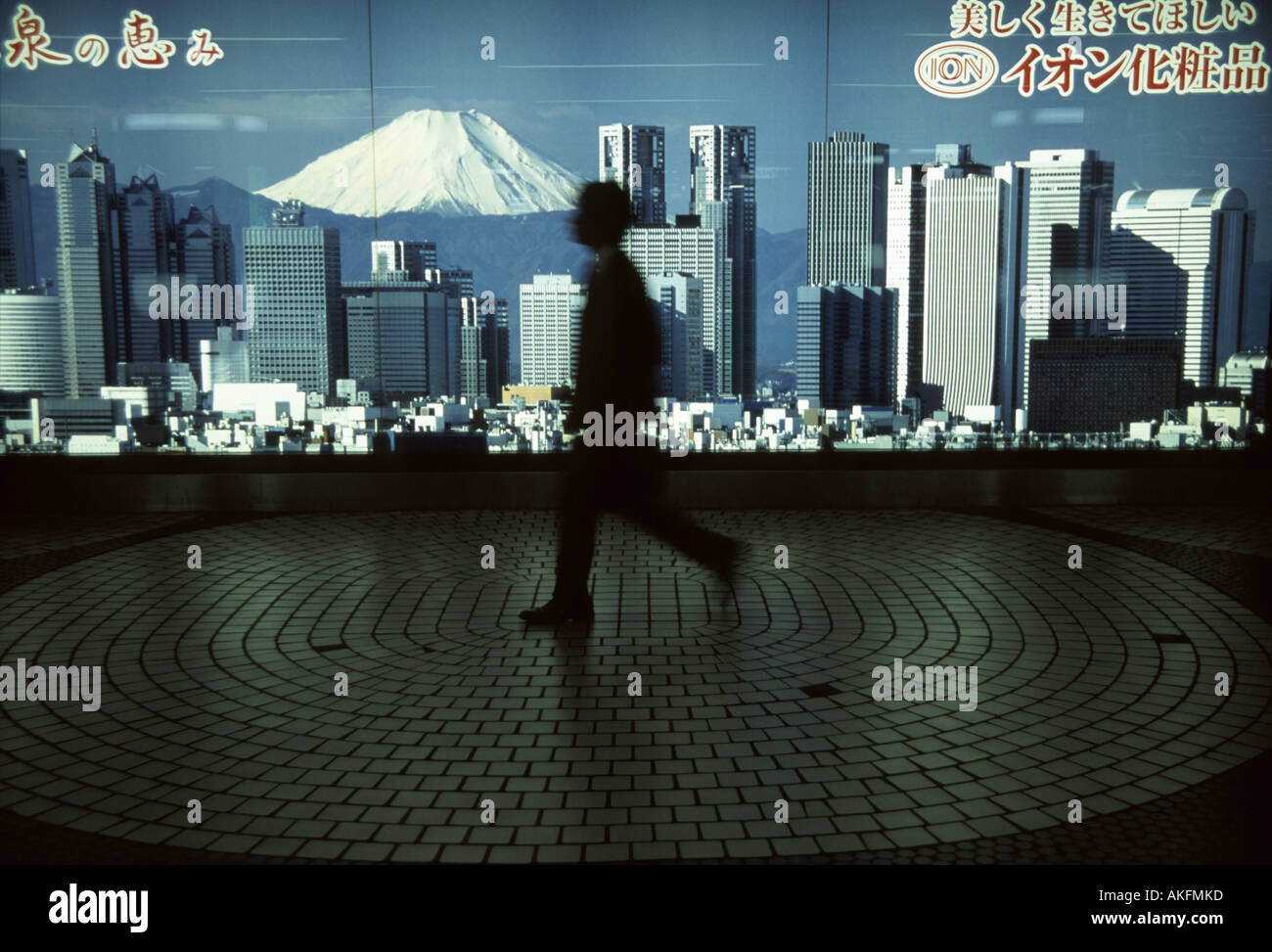 A Japanese salaryman walks past an advertising poster showing the Tokyo skyline and Mount Fuji in Shinjuku station, Tokyo, Japan Stock Photo