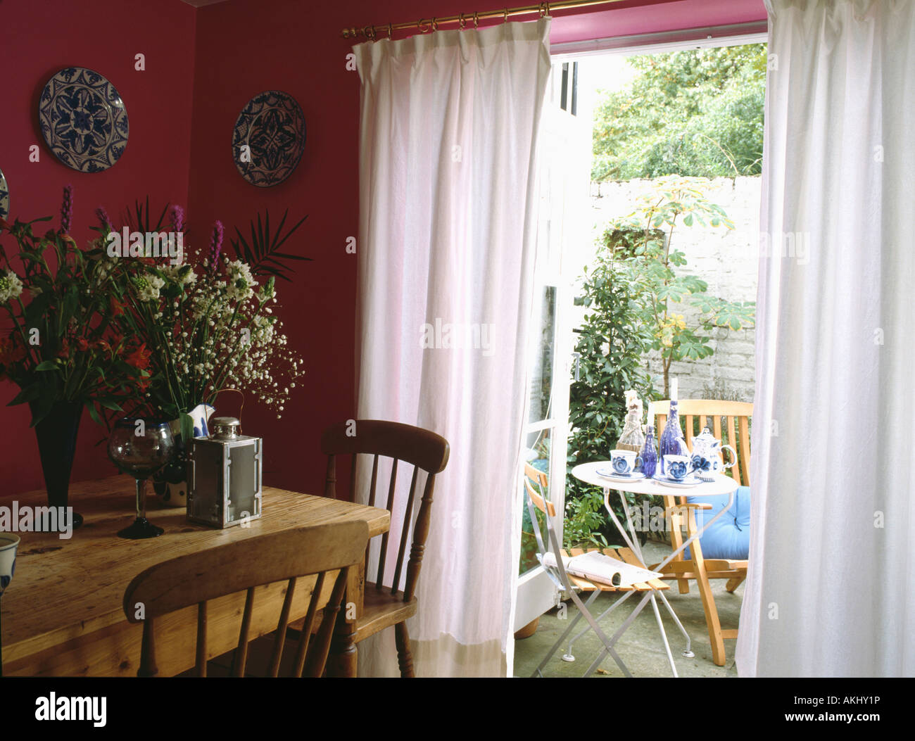 Pine table and chairs in pink diningroom with white curtains at French doors with view of patio garden Stock Photo