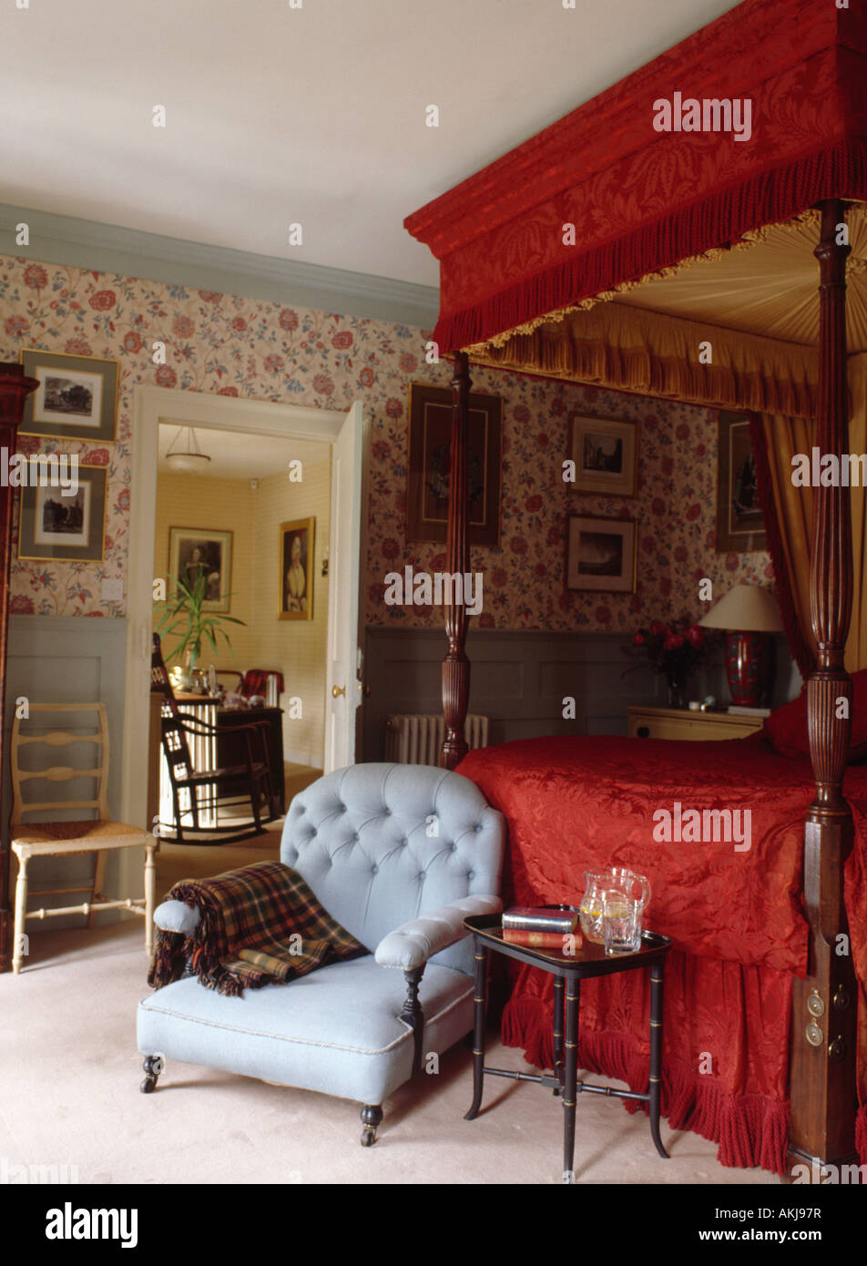 red silk canopy and cover on antique four poster bed in country bedroom with pastel blue armchair and floral wallpaper