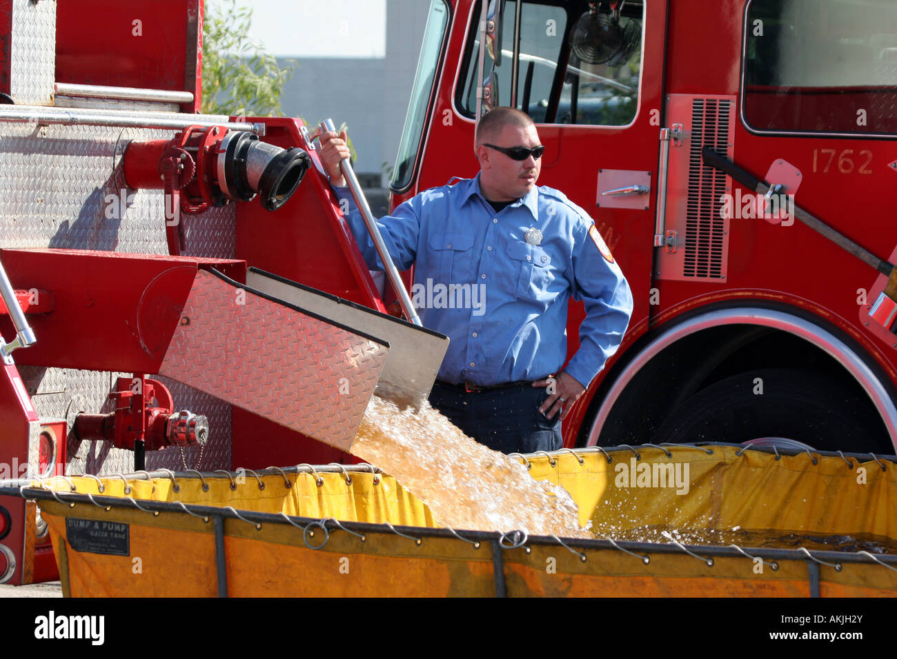 A firefighter standing at the unloading or dumping of water from a tranker fire truck into a dump and pump station Stock Photo