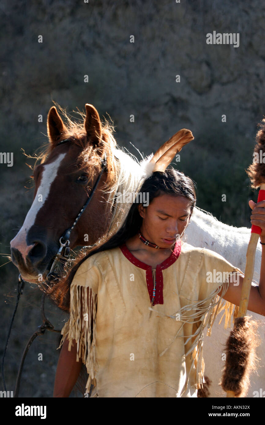 A Native American Indian boy wearing a feather standing next to a horse holding a spear with buffalo hide on it Stock Photo
