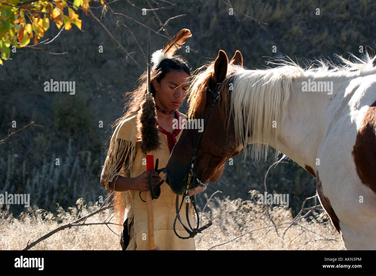 A Native American Indian boy wearing a feather standing next to a horse holding a spear with buffalo hide on it Stock Photo