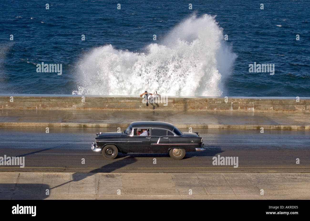 two young boys watching waves breaking over sea wall with old american car in foreground Stock Photo