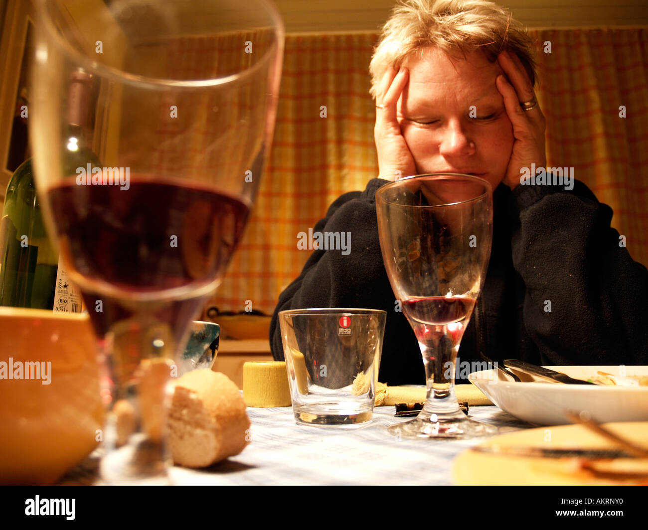 middle aged woman sitting after a meal at table holding her head in the hands in the foreground almost empty glasses of wine Stock Photo