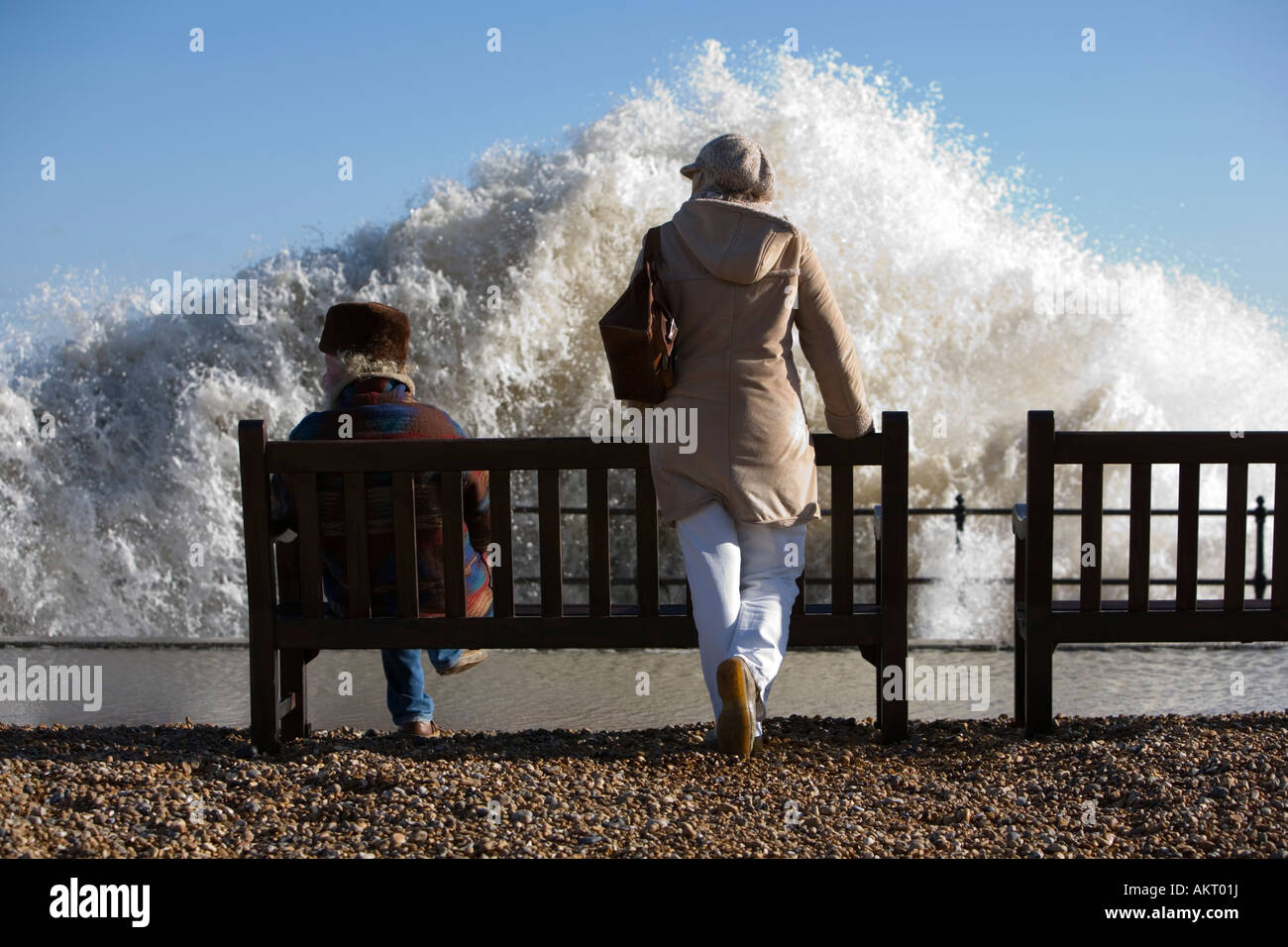 Couple on seafront benches as large wave breaks against sea wall Stock
