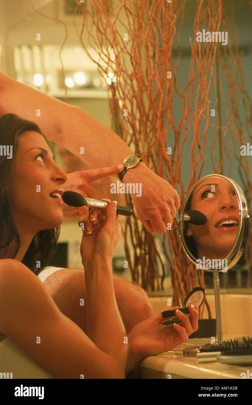 Woman putting on make up in bathroom with man pointing to watch showing time and patience Stock Photo
