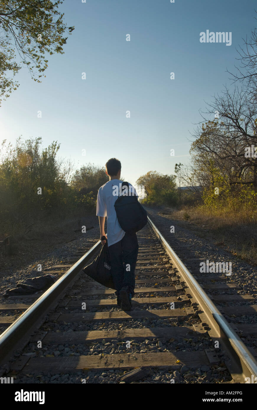 A 13-year-old boy walks down the center of a railroad track carrying his backpack and a sack of his belongings. USA. Stock Photo