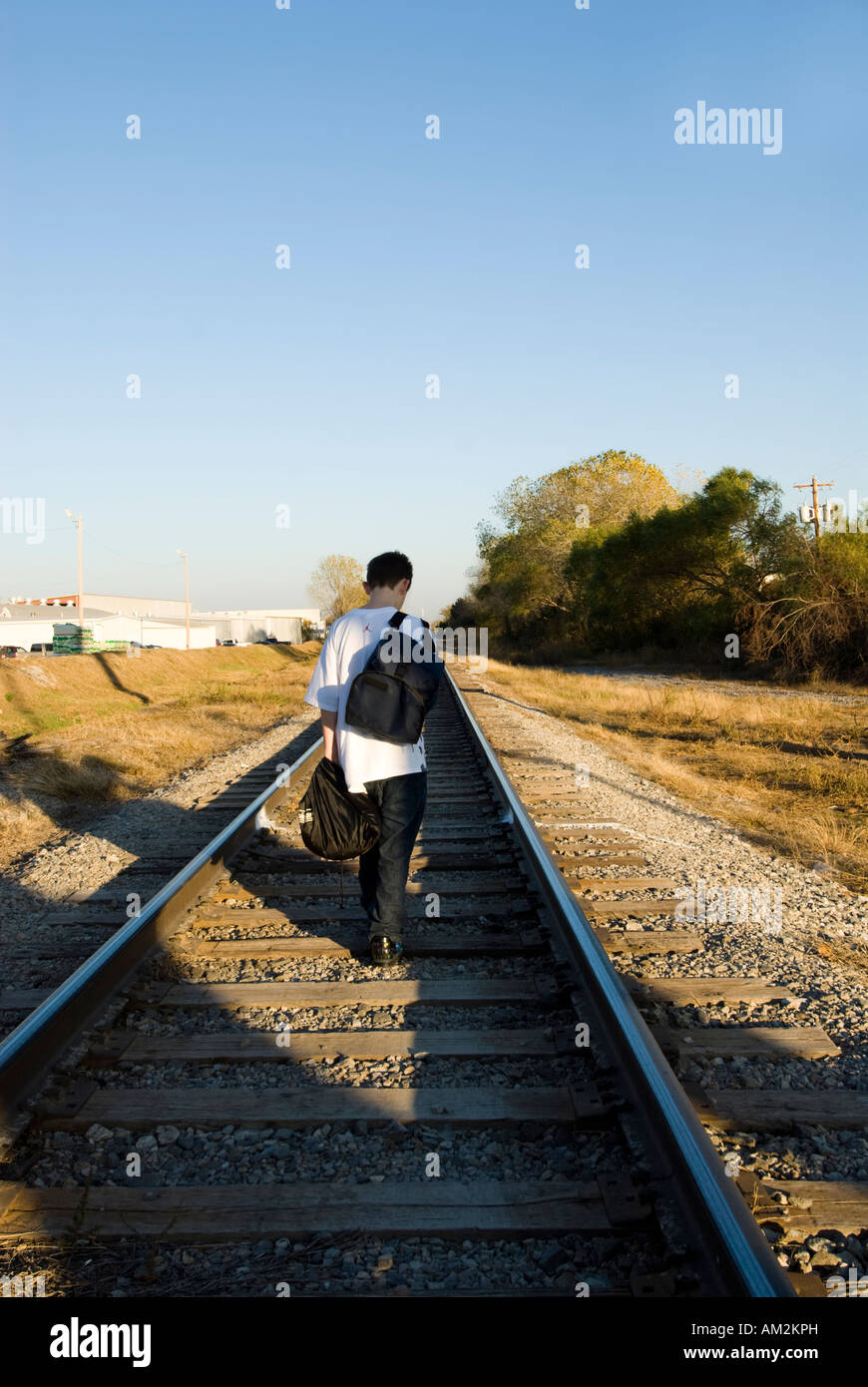 A 13-year-old boy walks down the center of a railroad track carrying his backpack and a sack of his belongings. Stock Photo