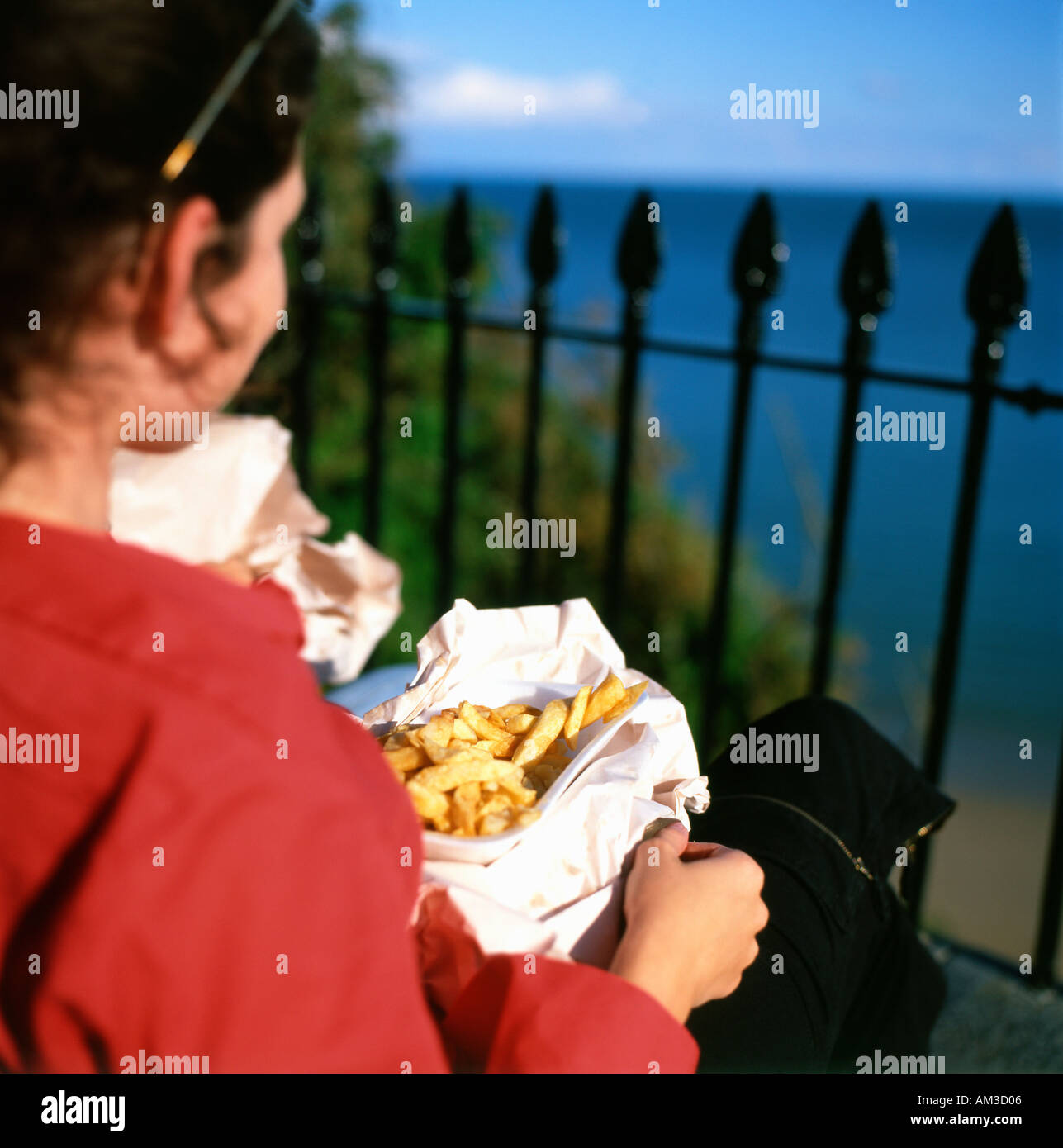 Rear back young view young woman eating fish & chips sitting above South Beach on the Welsh Coast Tenby Pembrokeshire Wales UK Britain  KATHY DEWITT Stock Photo