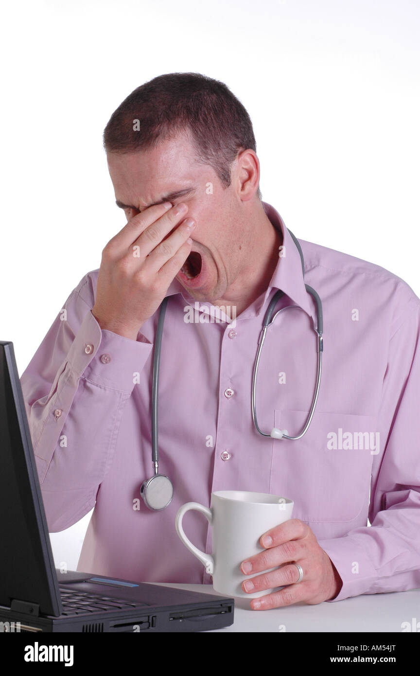 A young male doctor at his desk rubbing his eyes and yawning with fatigue from working long hours Stock Photo