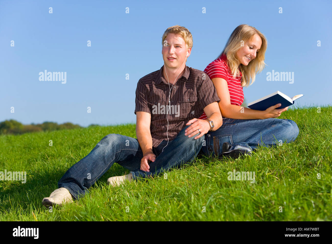 A young couple on a meadow, he is listening to music, she is reading a book Stock Photo