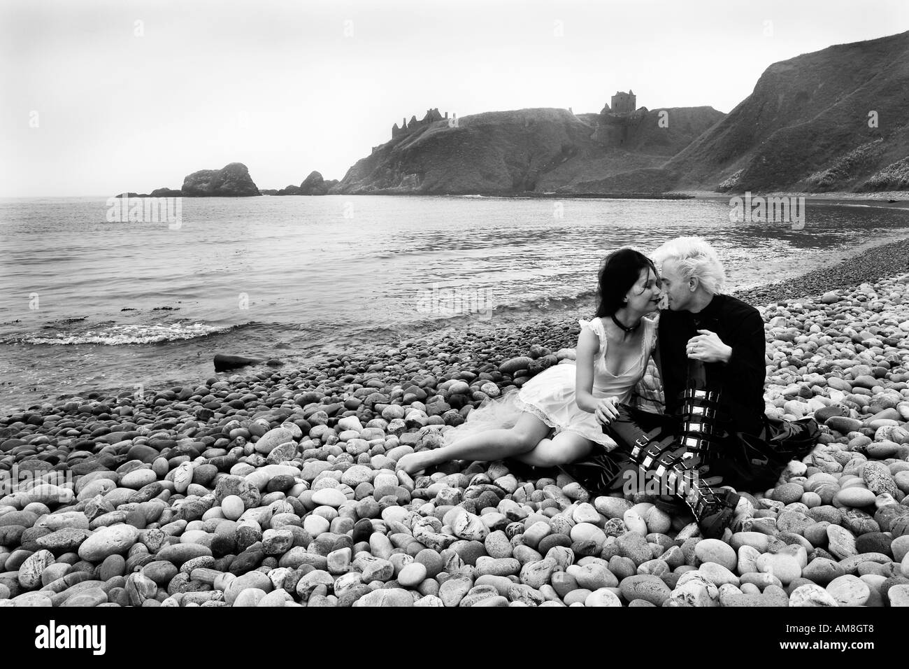A romantic gothic couple sitting on a beach with spectacular Dunnottar castle as a backdrop in black and white Stock Photo
