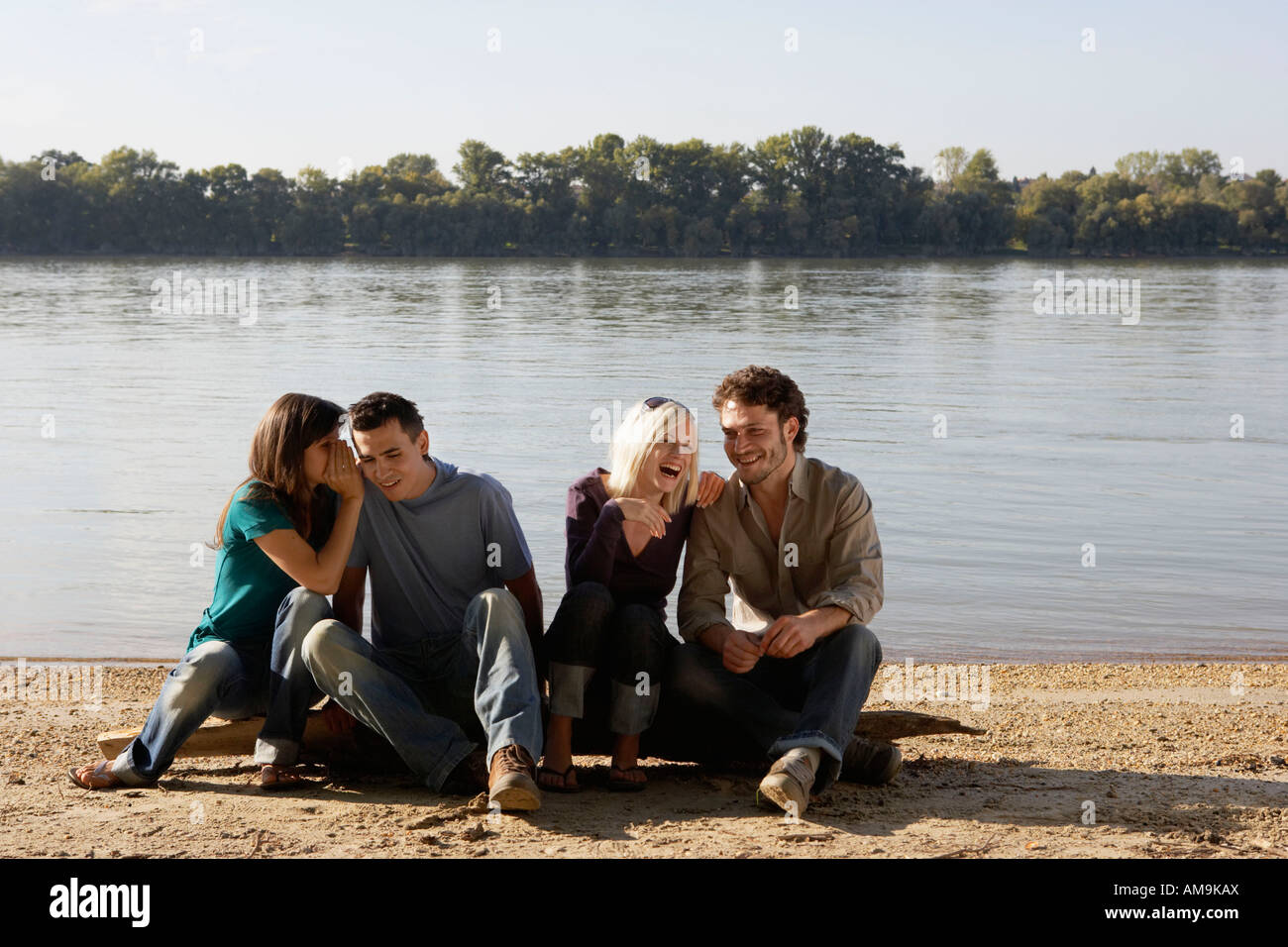 Four friends sitting on log by a lake smiling. Stock Photo