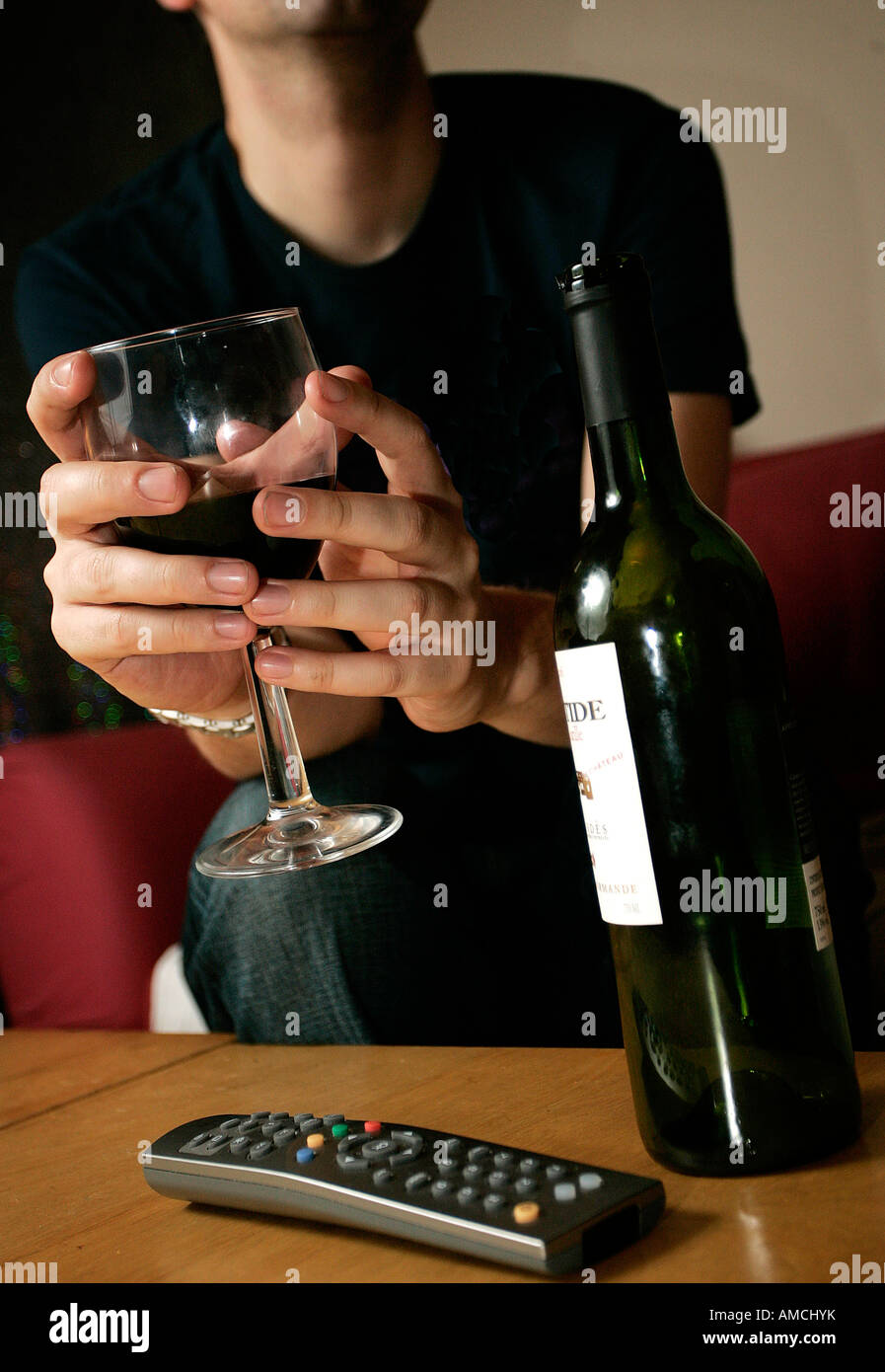 A young man enjoys a glass of wine at home. A television remote control sits on the table in front of him. Stock Photo