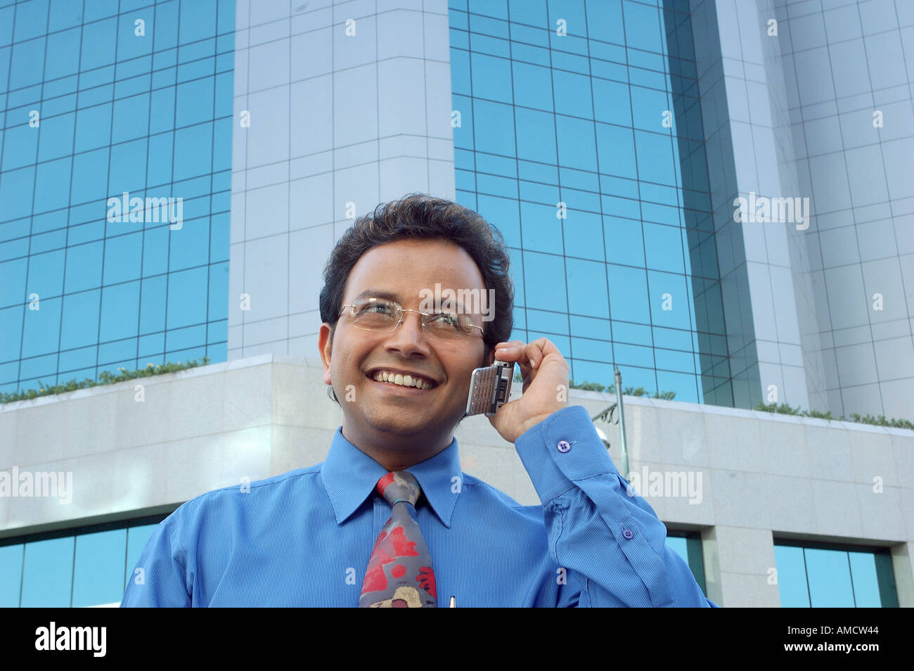 VEP72167 Young Indian Executive with spectacles smiling and talking on cell phone in front of modern building Stock Photo