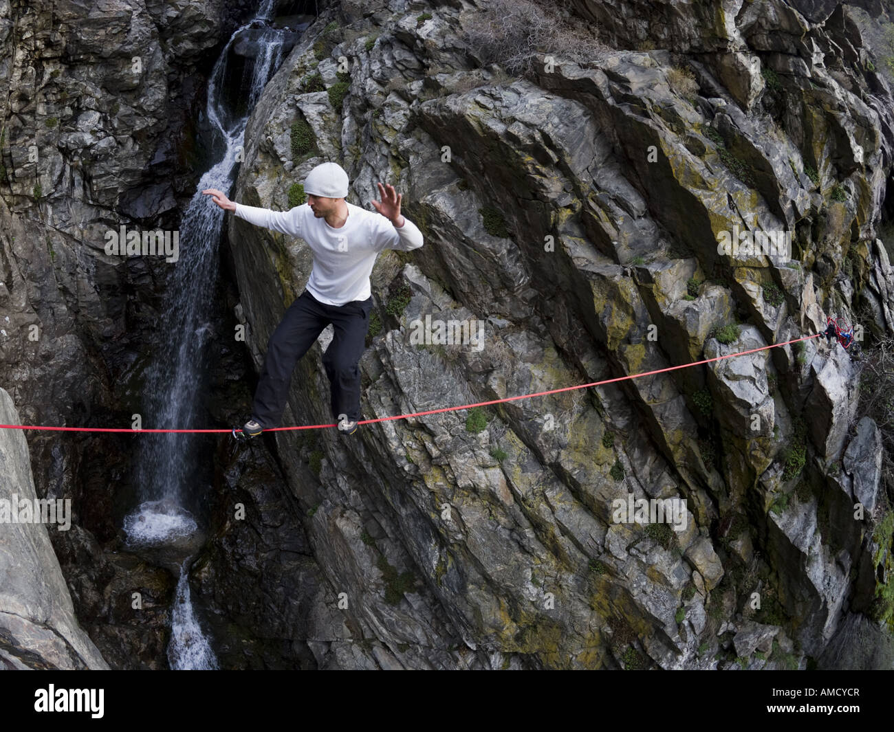 Man walking across rope with rock and waterfall in background Stock Photo