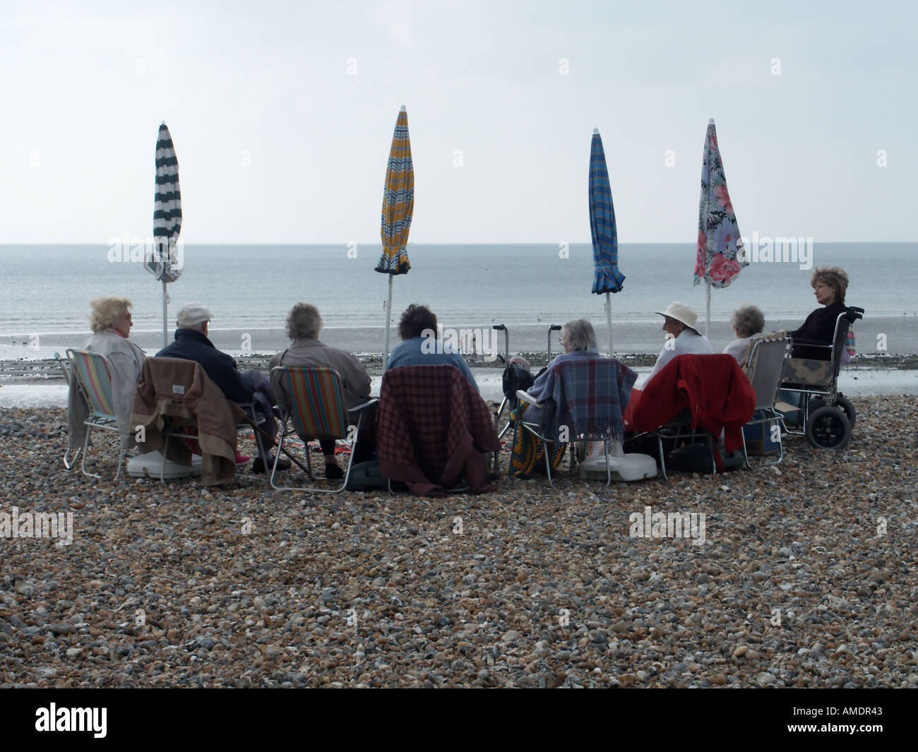 A group of senior citizens on a day out sitting together on pebble stony beach anticipating some sun sunshade ready or perhaps rain Sussex England UK Stock Photo