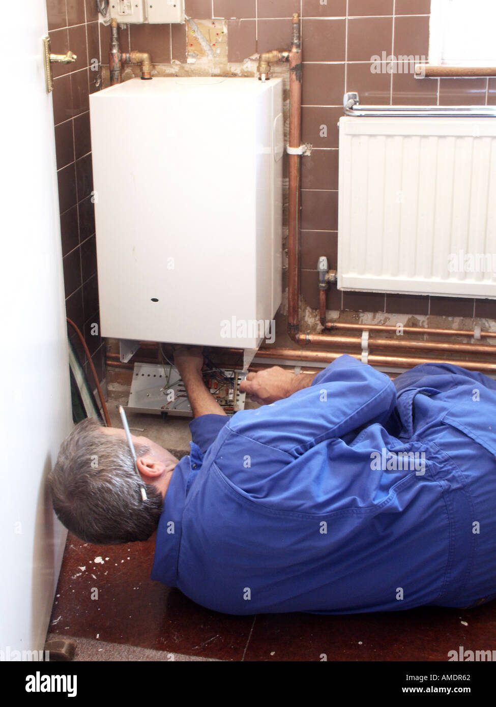 Back view plumber & heating engineer working in house interior on gas central heating boiler checking copper pipework & controls in house England Uk Stock Photo