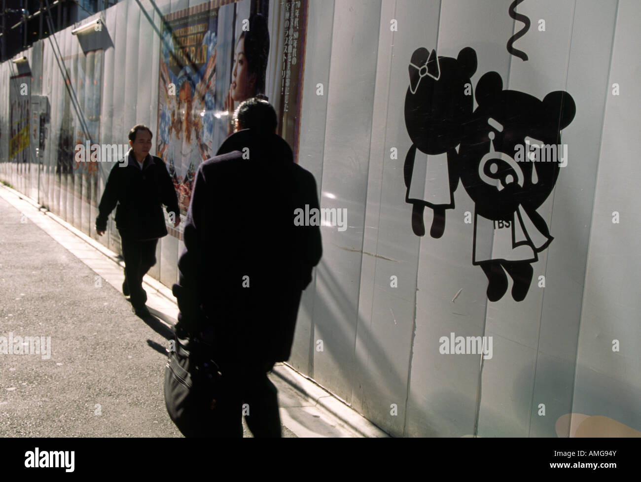 Two men walking in a Tokyo street with an image of a cartoon bear crying on a construction site wall to their side. Tokyo, Japan Stock Photo