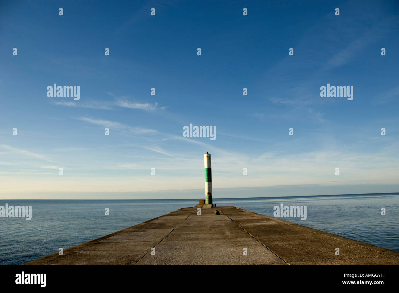 green and white striped lighthouse tower at end of a concrete jetty pointing out to sea on a calm clear blue sky day Stock Photo