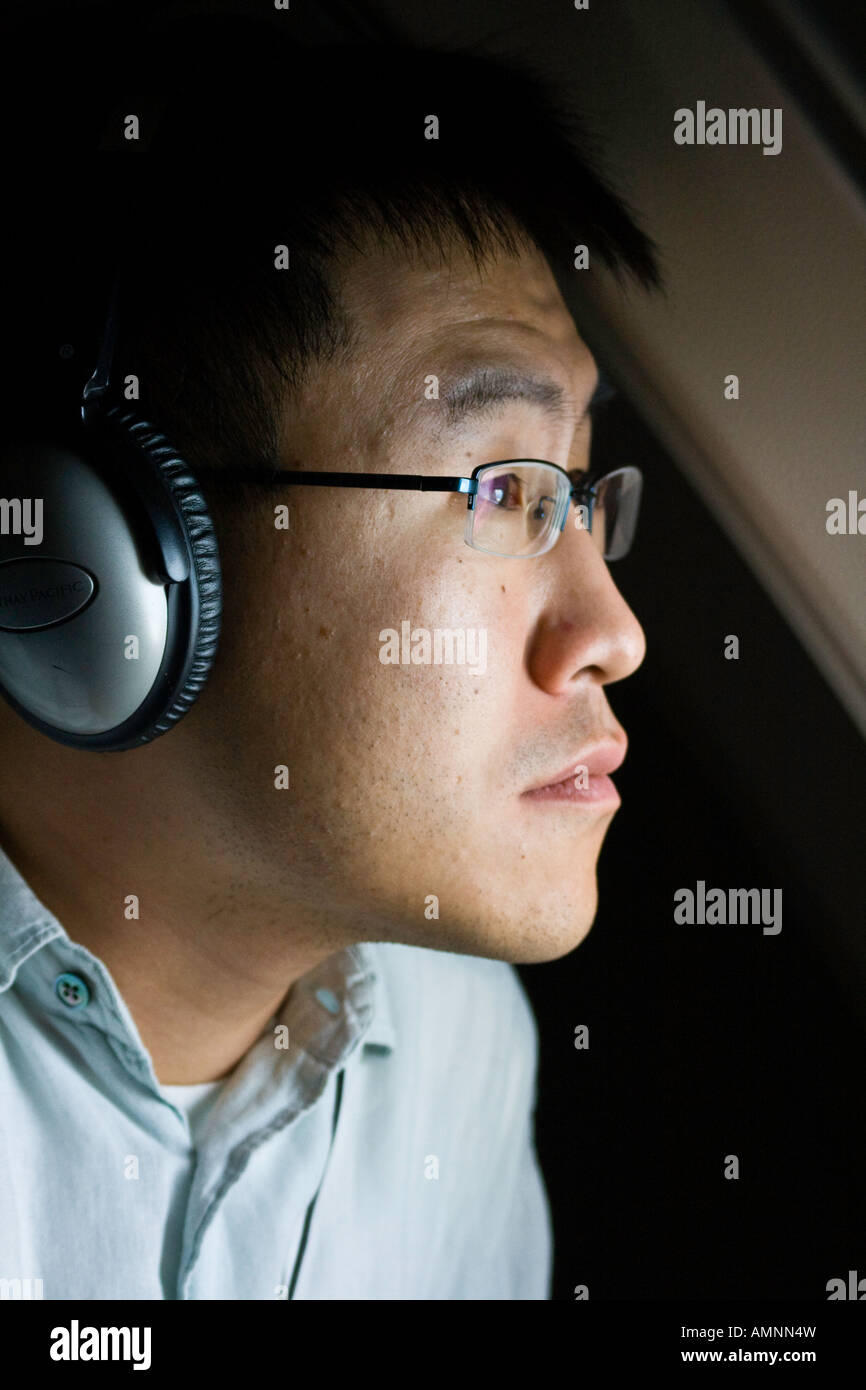 Young Korean American Man Passenger with Headphones on and Looking out the Window of a Commercial Plane Stock Photo
