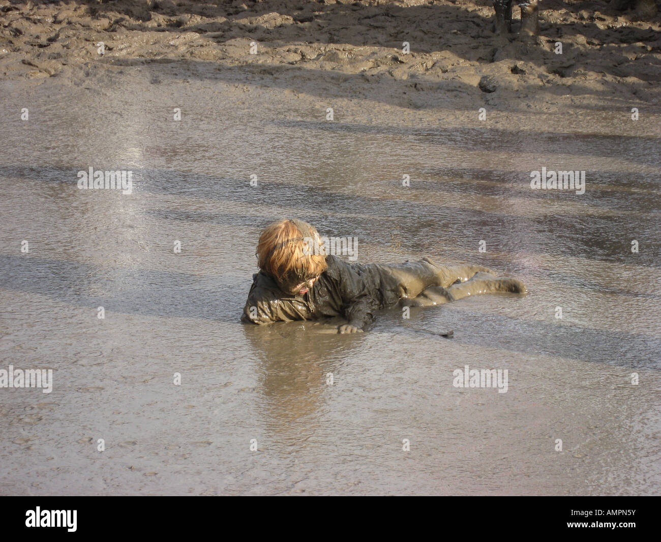 Young boy covered completely in mud lying in large mud pond at