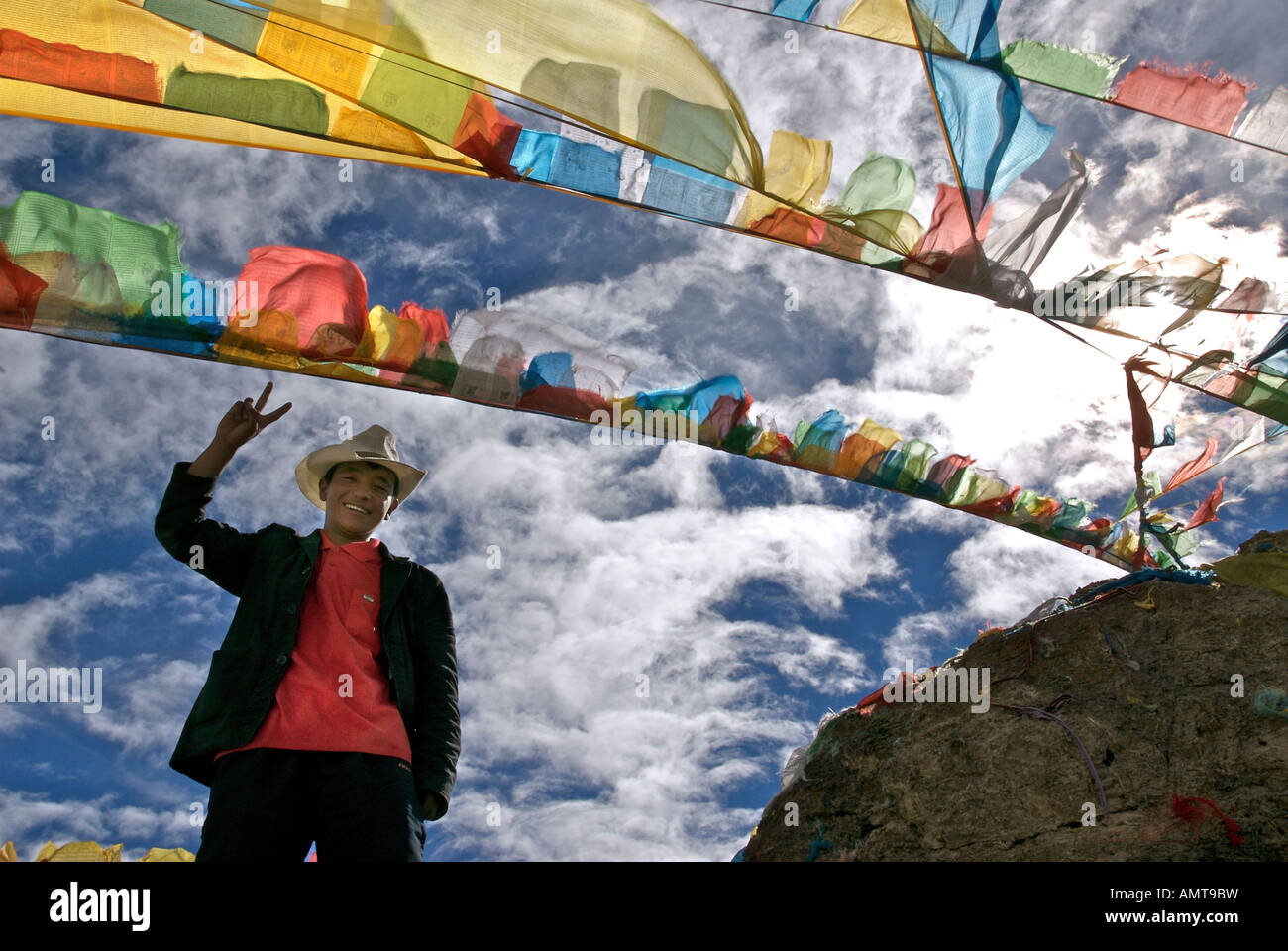 Tibetan man in a cowboy hat making a peace sign beneath prayer flags NamTso lake Tibet Stock Photo